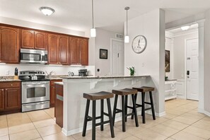 Cozinha com armários de madeira, eletrodomésticos de aço inoxidável, ilha central com banquetas, iluminação pendente e relógio na parede.
