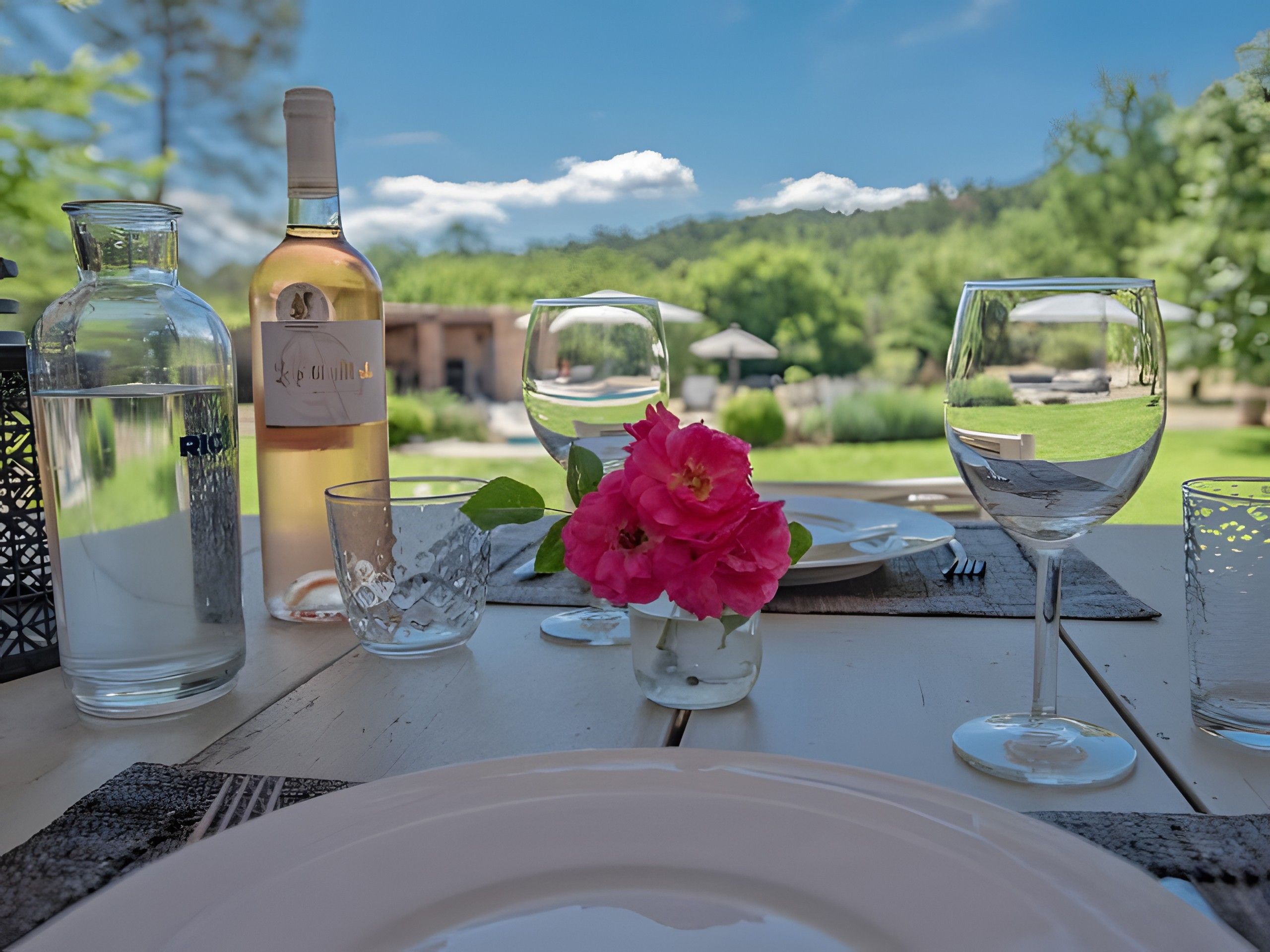 A table set outdoors with a bottle of wine, glasses, and a flower centerpiece.