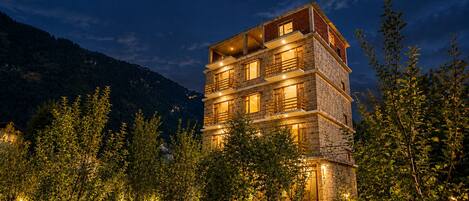 A multi-story building with balconies, surrounded by trees and a mountainous backdrop.