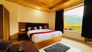 Een slaapkamer met een houten plafond, een bed met wit beddengoed en rode accenten, een nachtkastje, een afstandsbediening op een bruine tafel en uitzicht op groen buiten.