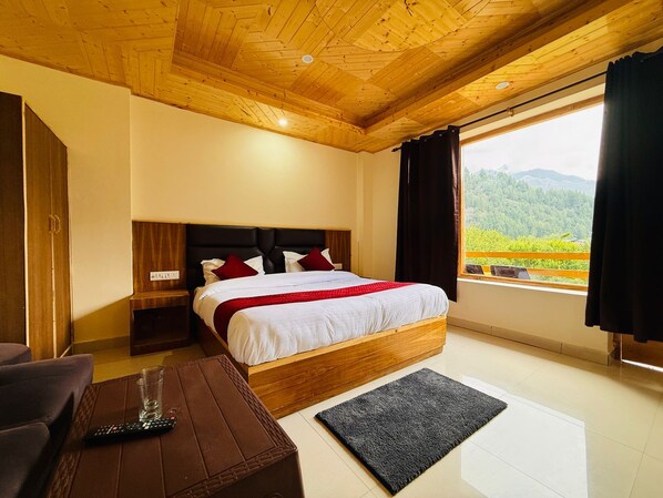 Un dormitorio con techo de madera, una cama con ropa de cama blanca y detalles rojos, una mesita de noche, un control remoto sobre una mesa marrón y vistas a zonas verdes al exterior.