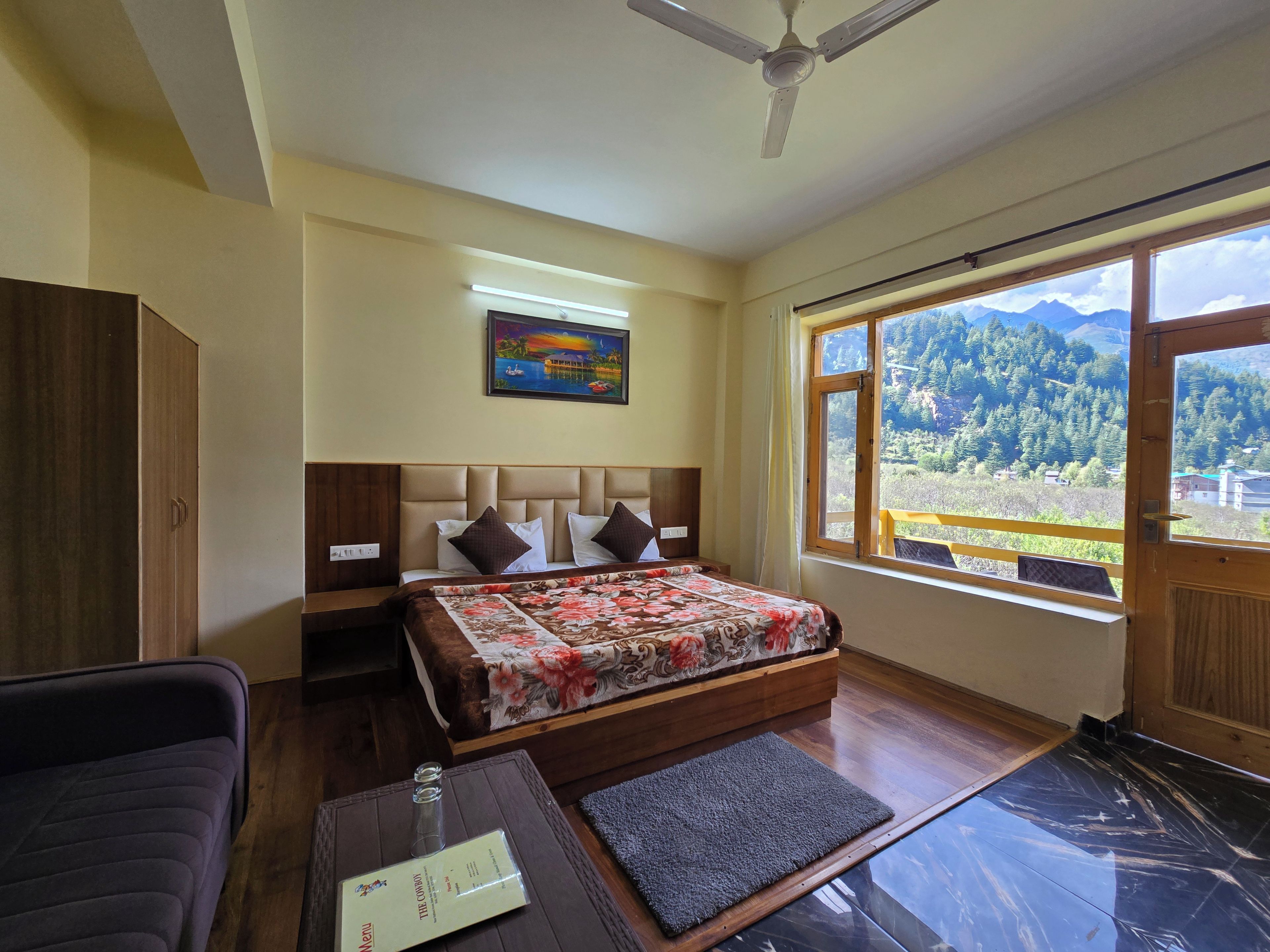 Une chambre à coucher comprenant un lit, une table de chevet, une armoire, un bureau avec une chaise, une fenêtre donnant sur les montagnes et un ventilateur de plafond.