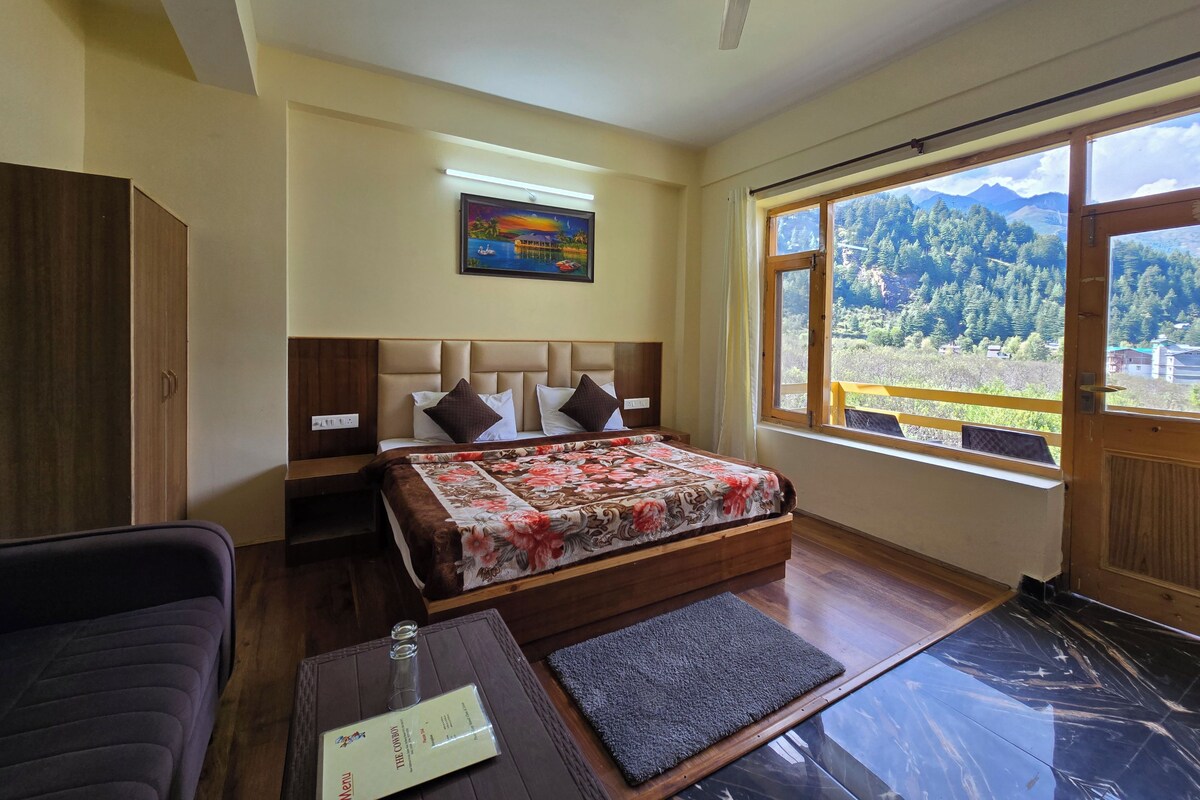 A bedroom with a bed, a nightstand, a wardrobe, a desk with a chair, a window with a view of mountains, and a ceiling fan.