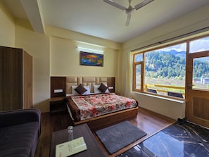 A bedroom with a bed, a nightstand, a wardrobe, a desk with a chair, a window with a view of mountains, and a ceiling fan. - The Cow Boy Manali (Manali)