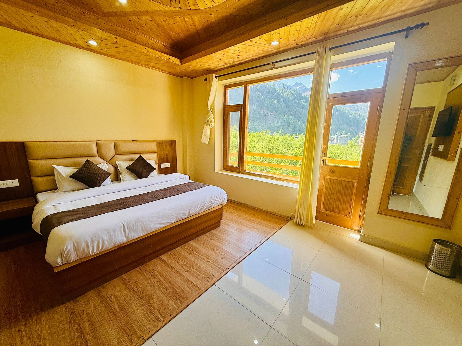 Un dormitorio con cabecera de madera, una cama grande con ropa de cama blanca, una ventana con vistas a zonas verdes y un espejo en la pared.