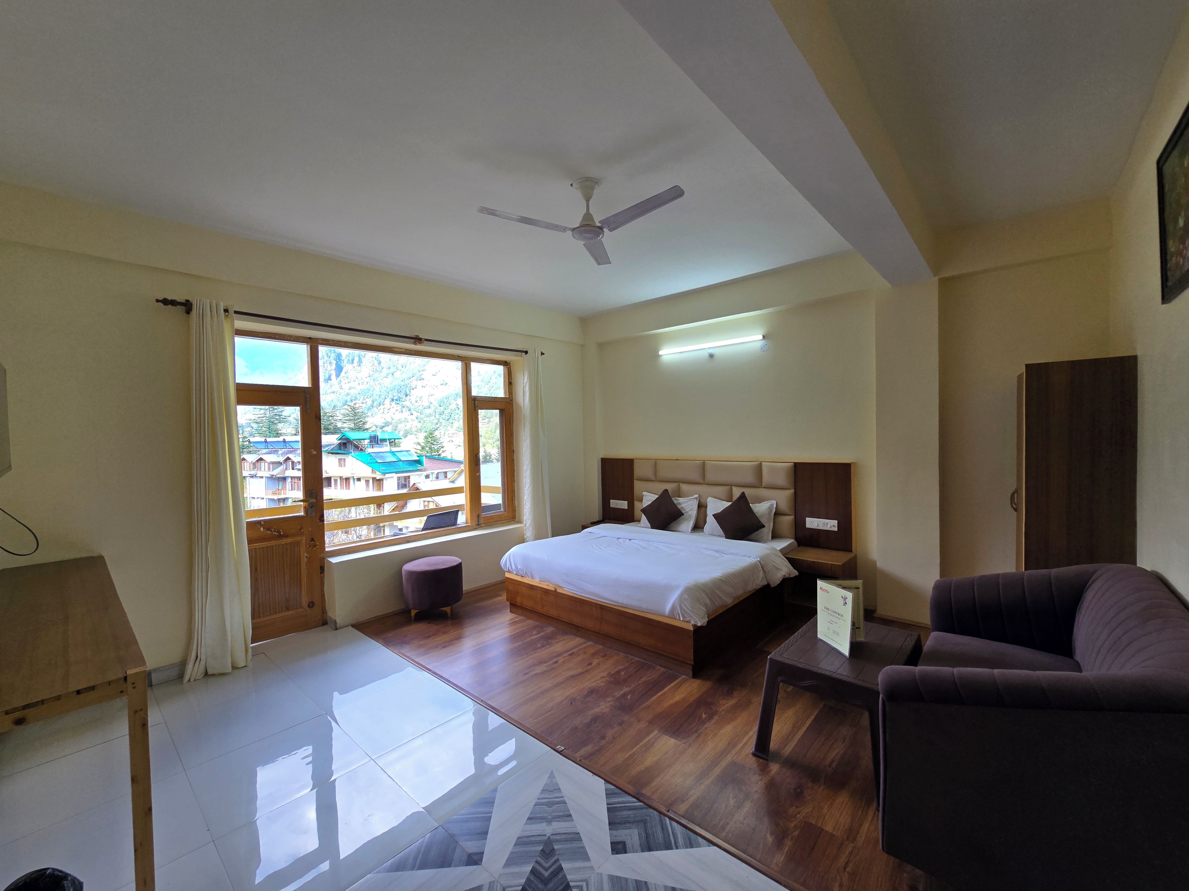 Une chambre d’hôtel comprenant un lit, un canapé, un bureau, une fenêtre avec des rideaux, un ventilateur de plafond et une vue sur les bâtiments environnants.