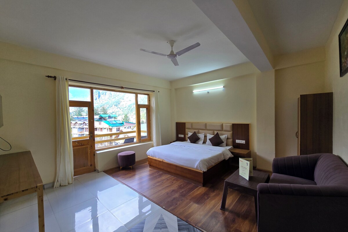 A hotel room with a bed, a sofa, a desk, a window with curtains, a ceiling fan, and a view of buildings outside.