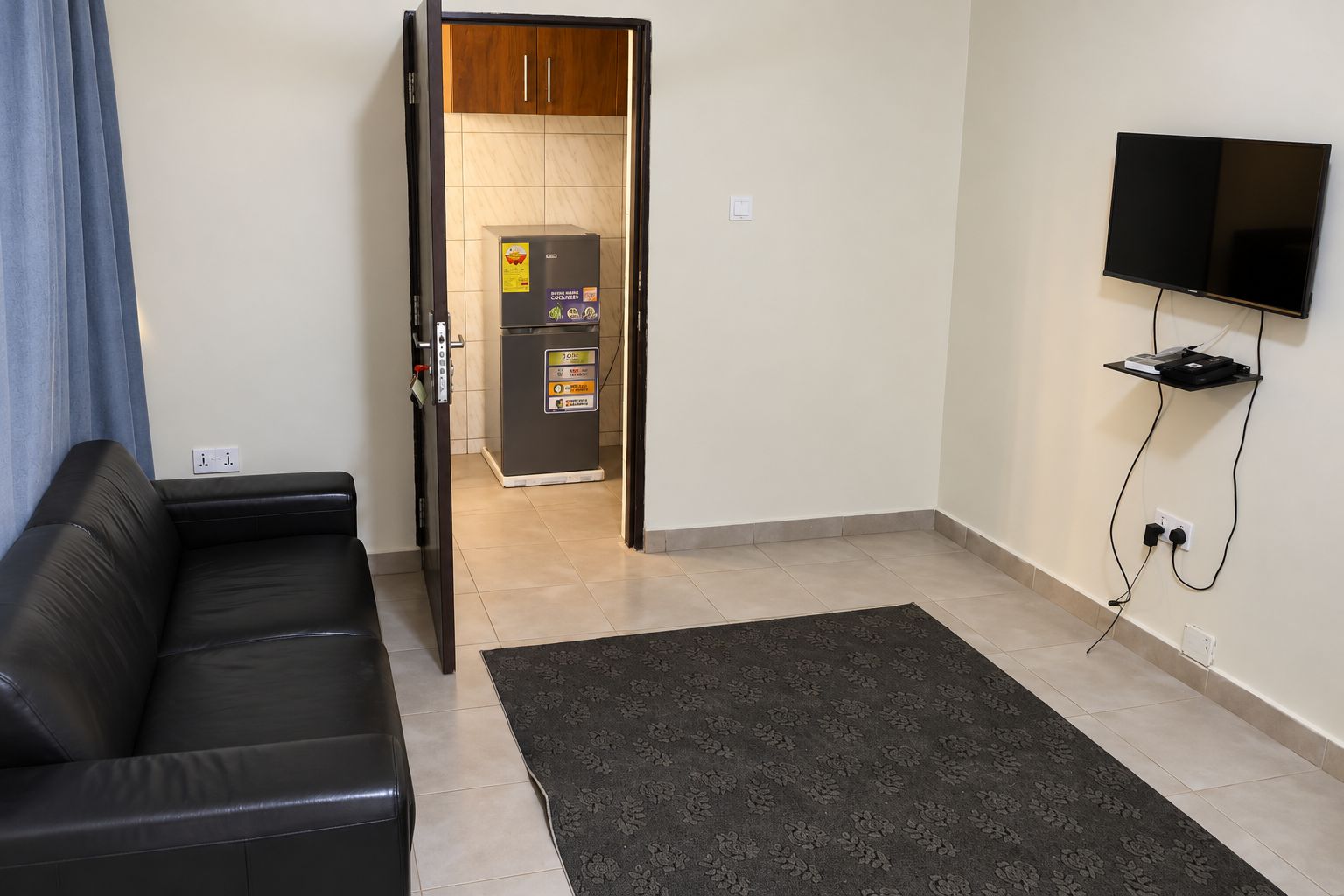 A living room with a black leather sofa, a dark patterned rug, a television mounted on the wall, and a kitchen area visible through an open door.