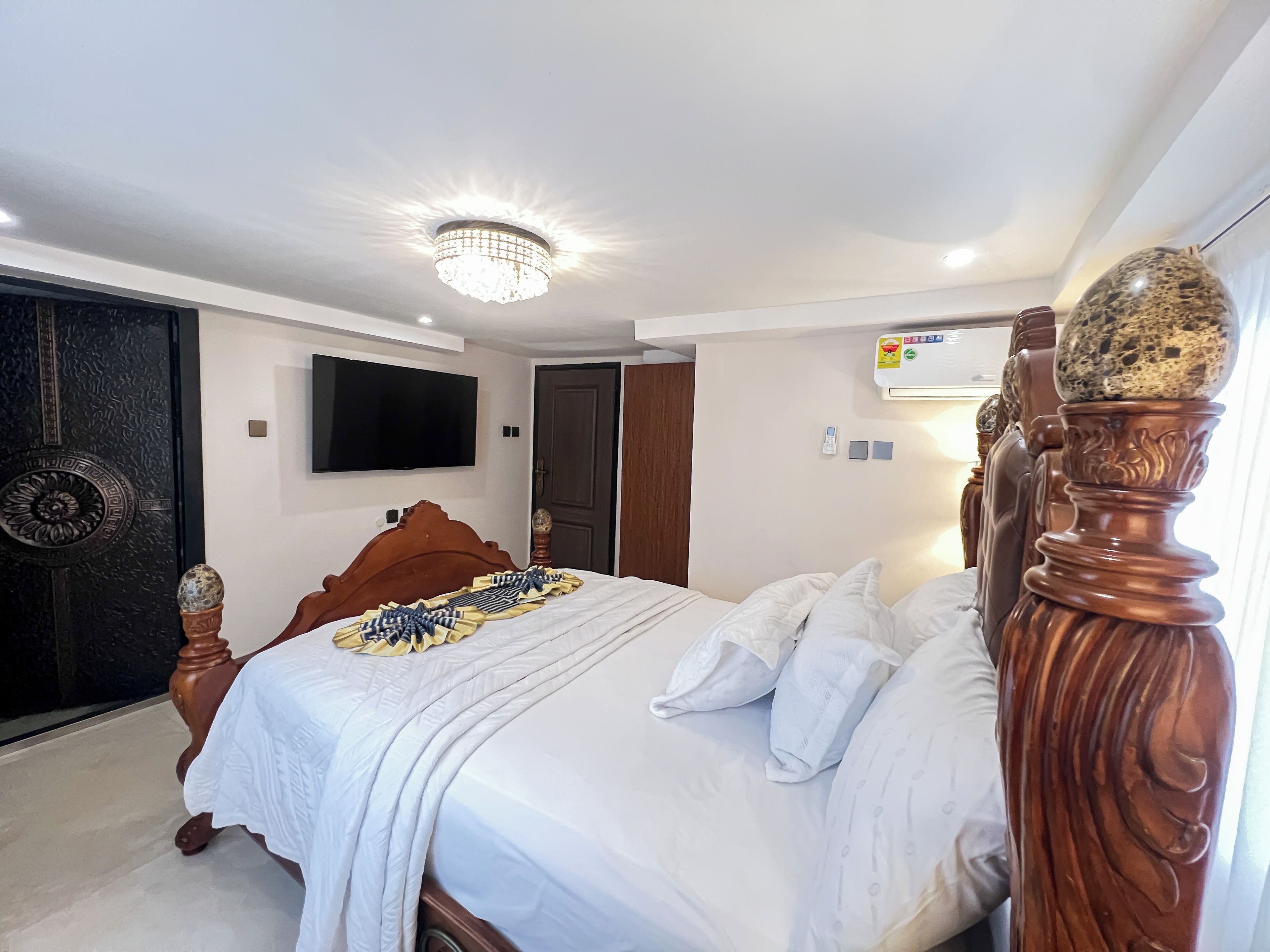 A neatly made bed with a wooden headboard, a television mounted on the wall, and a ceiling light fixture.