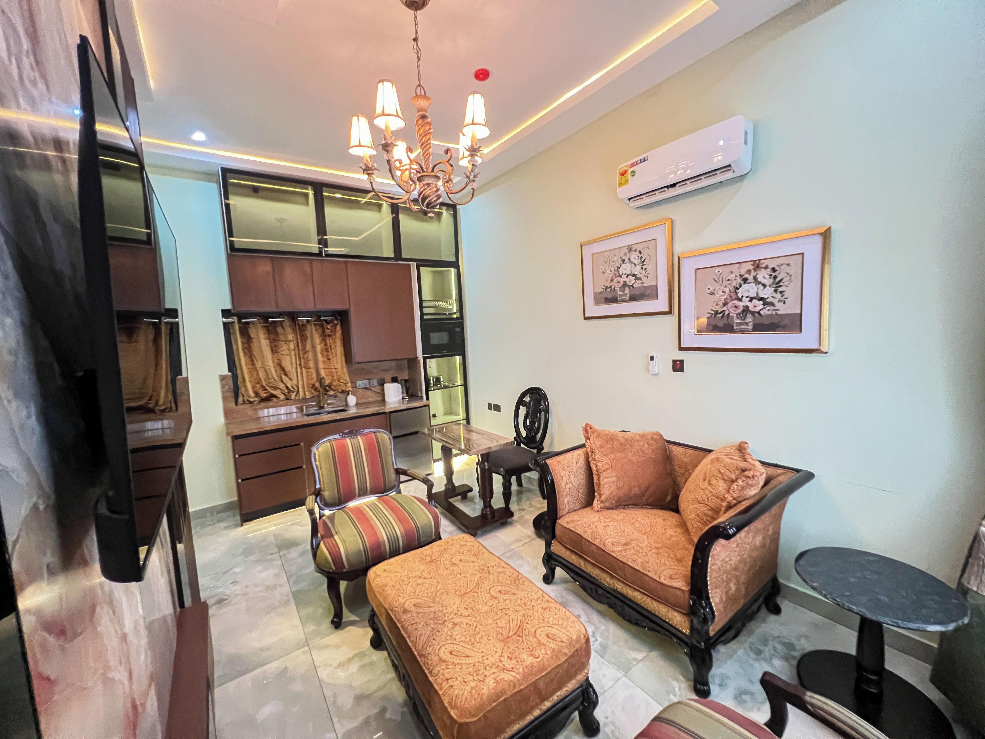 A living room with a sofa, ottoman, and a small table. There is a kitchen area with cabinets and a window with curtains.