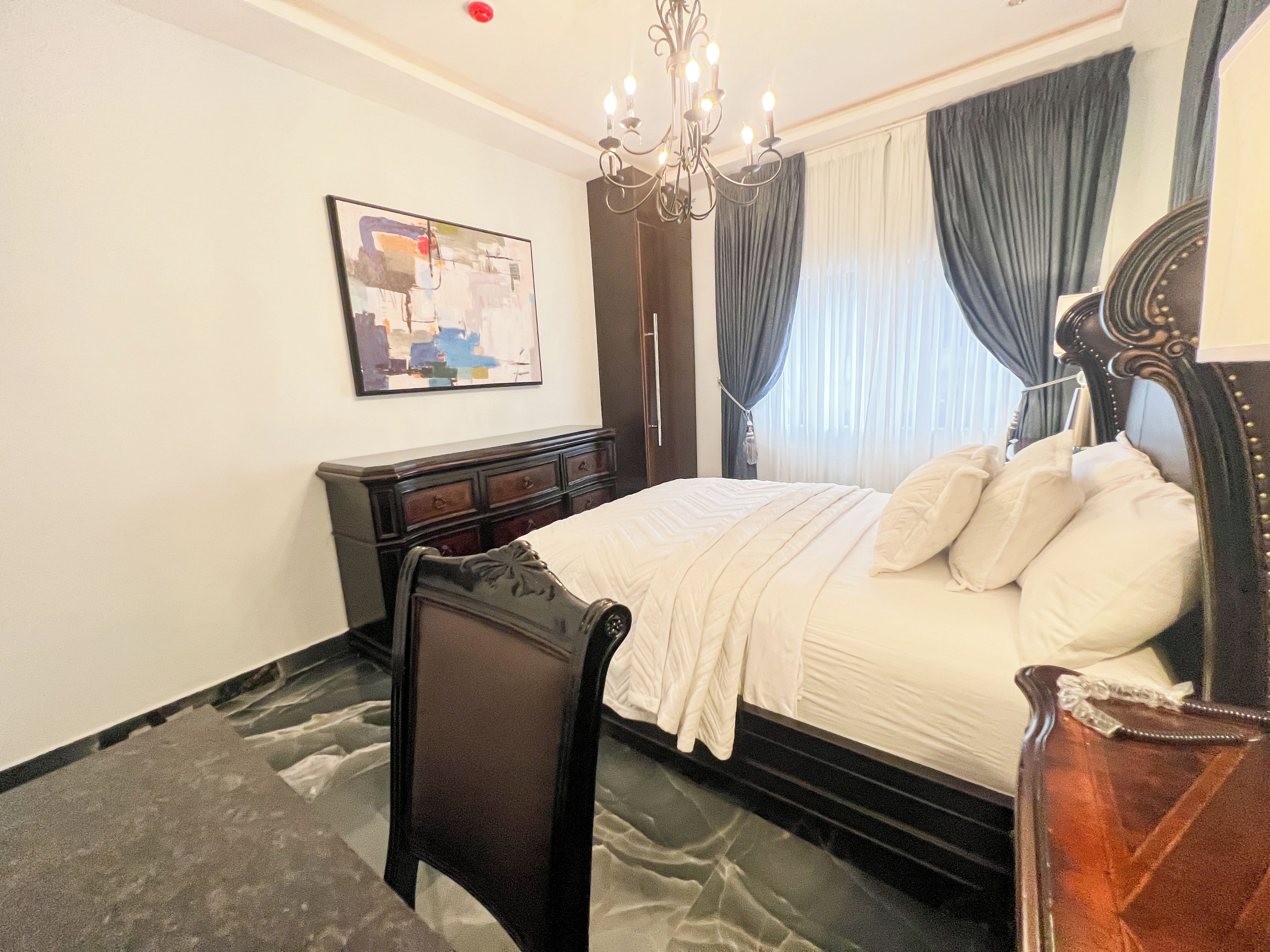 Une chambre d’hôtel avec un lit, une chaise, une commode, un lustre et un tableau au mur.