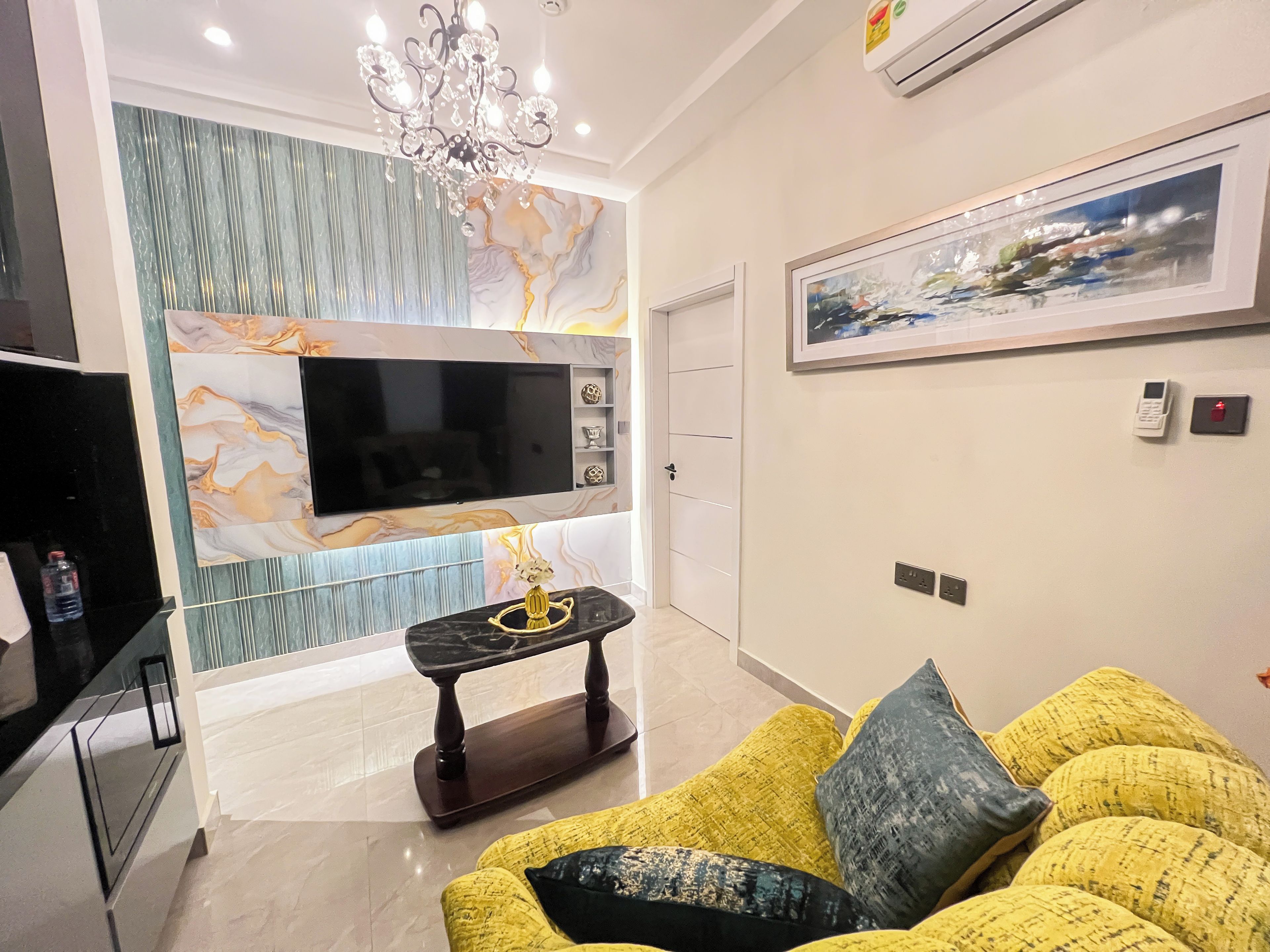 Ein modernes Wohnzimmer mit Flachbildfernseher, Wand im Marmor-Design, einer Kristallleuchtre und einem gelben Sofa mit Kissen.