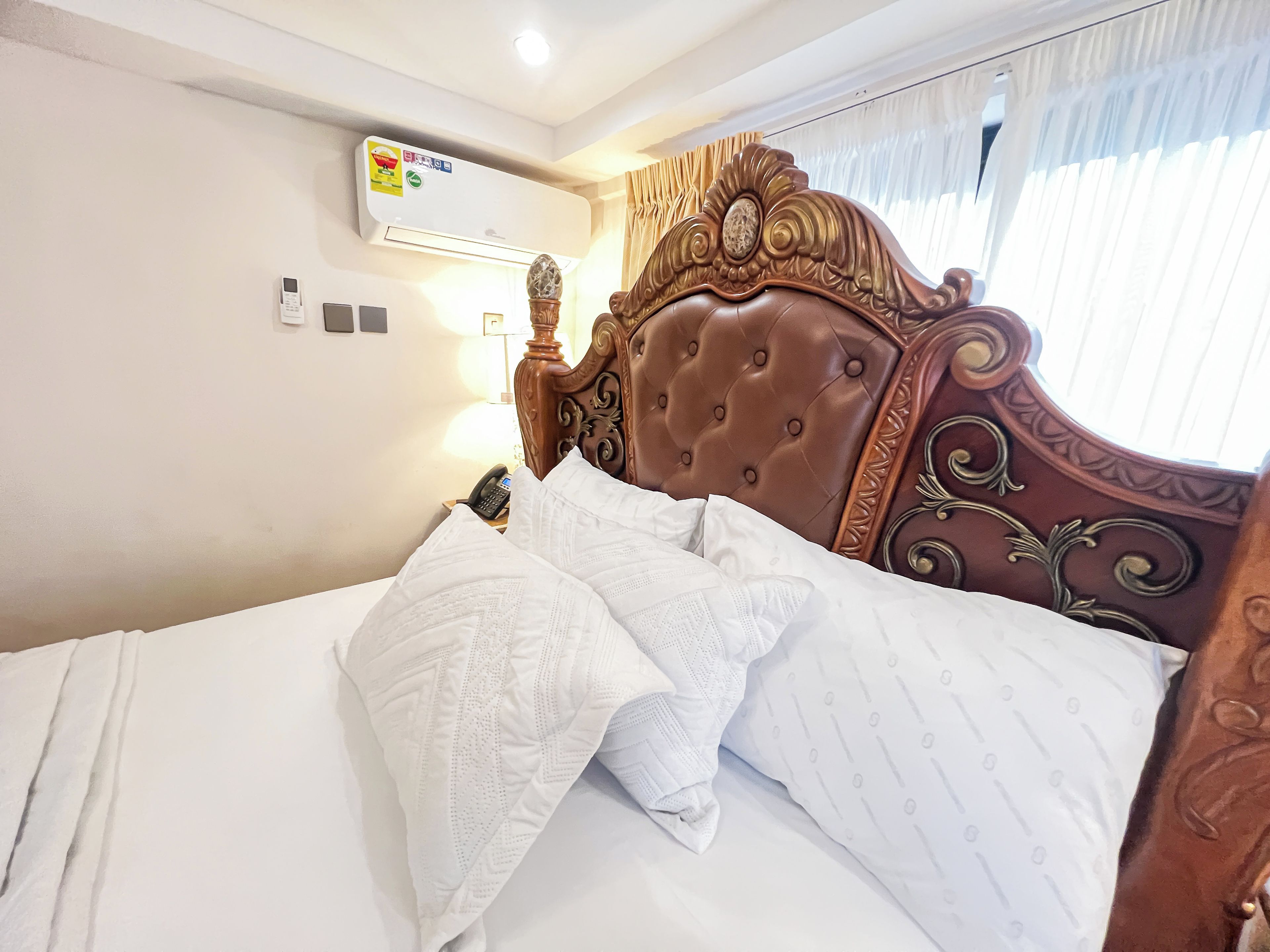 A hotel room with a bed, a bedside telephone, an air conditioning unit, and a window with curtains.
