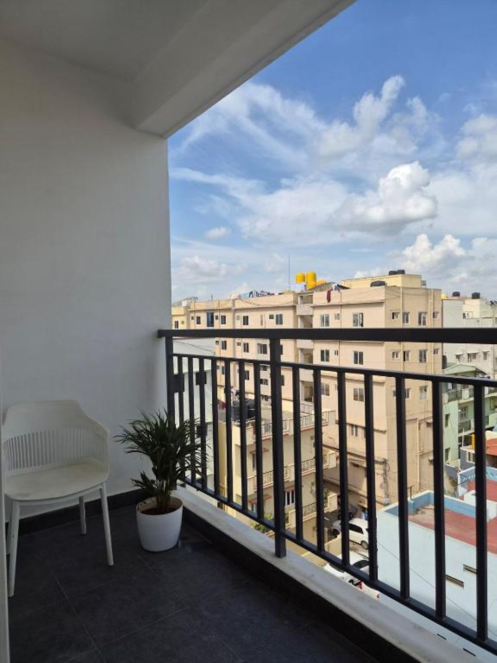 A balcony with a white chair and a potted plant, overlooking a residential area with multiple buildings.