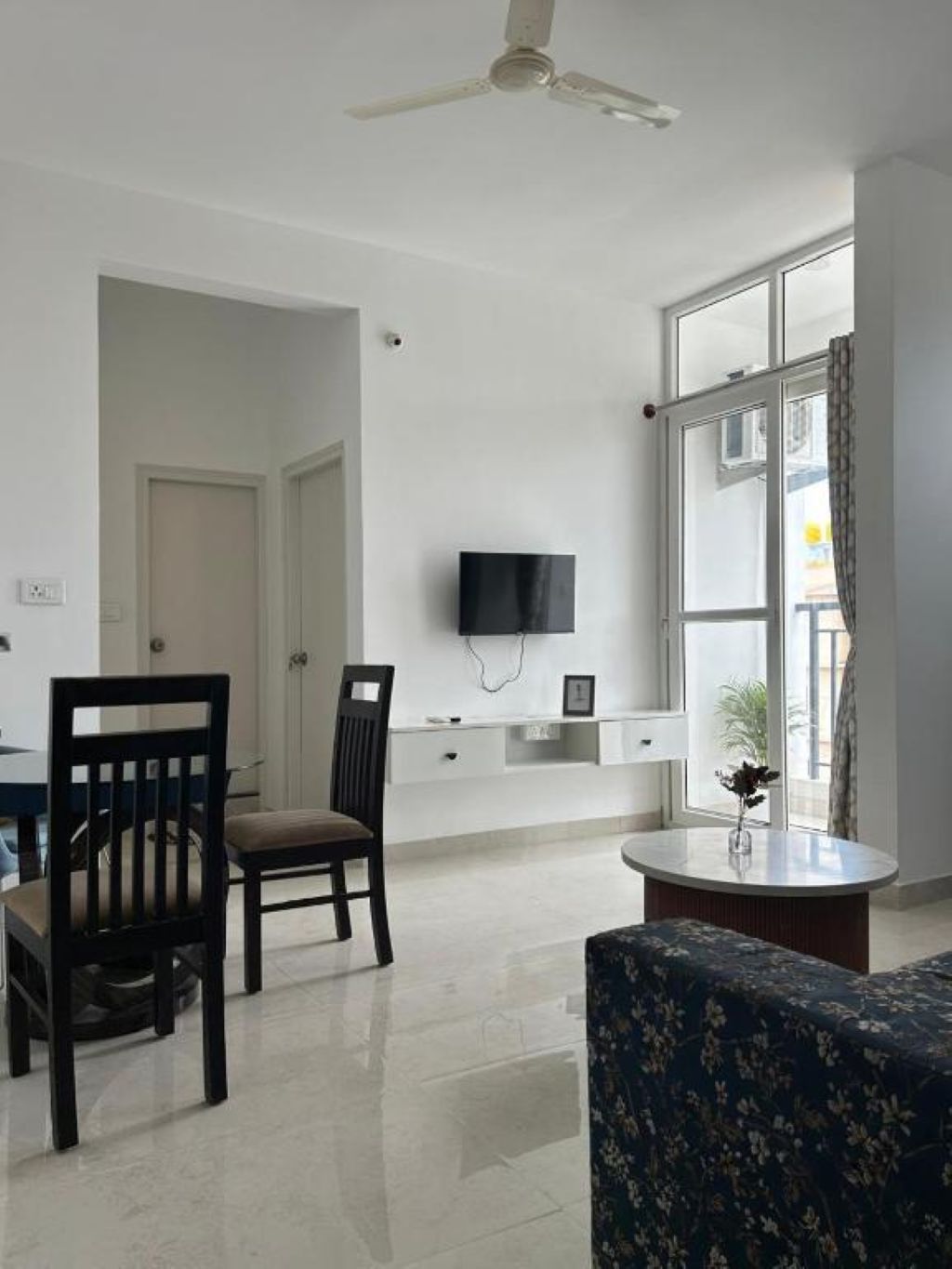 A modern living room with a dining table, two chairs, a sofa, a ceiling fan, a flat-screen TV mounted on the wall, and a large window with curtains.
