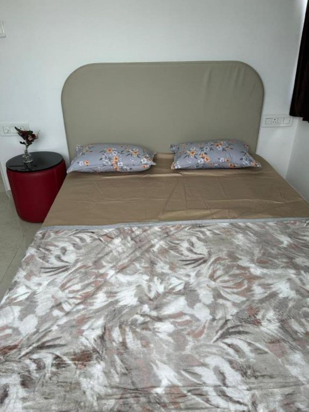 A bedroom with a bed, two pillows, a red side table, and a vase with flowers.