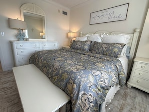 A bedroom with a bed, a bench, a dresser, a mirror, and a wall sign. - Gulf & Pool Views • Private Beach Access • Walk to Shops & Restaurants in Marco (Marco Island)
