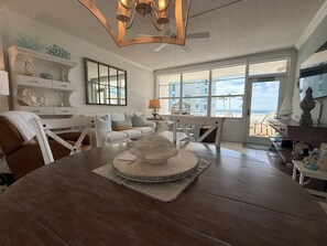 A spacious living room with a large wooden dining table, comfortable seating, a decorative centerpiece, and a view of the ocean through large windows. - Gulf & Pool Views • Private Beach Access • Walk to Shops & Restaurants in Marco (Marco Island)