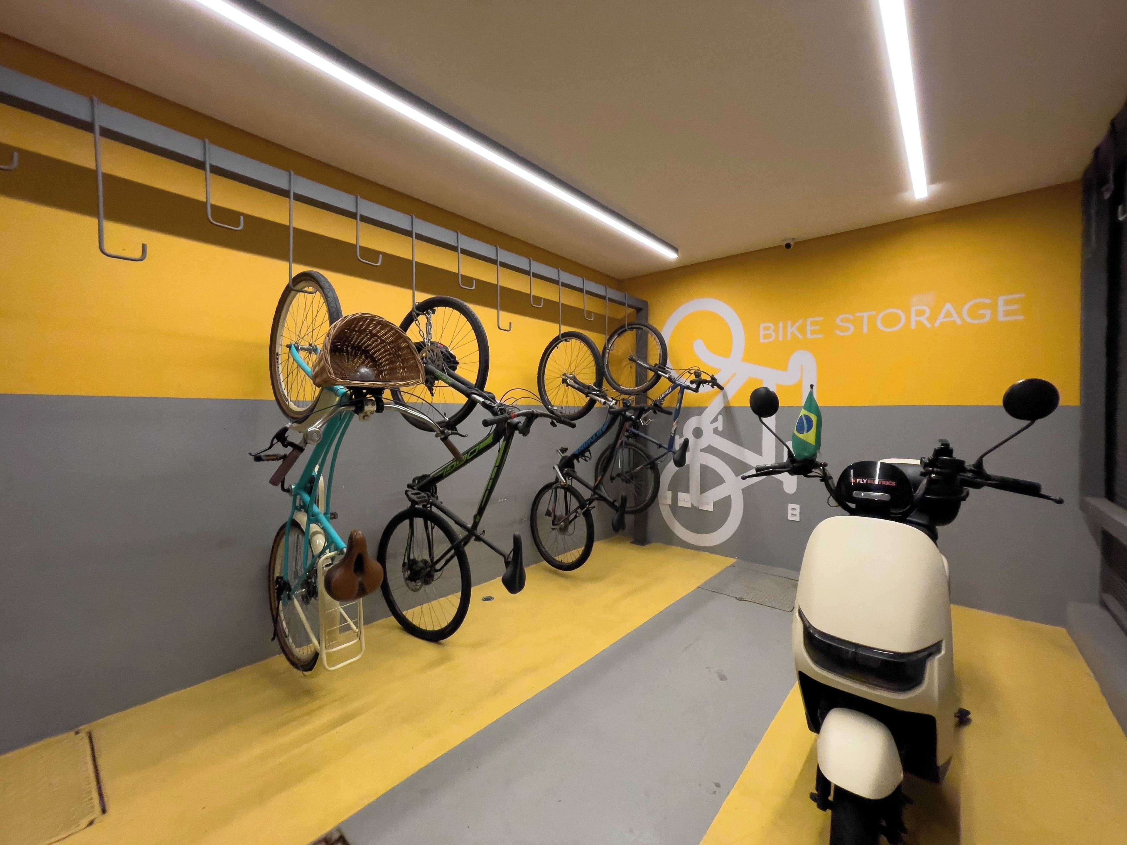 A bike storage area with multiple bicycles and a scooter, featuring a yellow and grey color scheme and a wall with a bike storage sign.