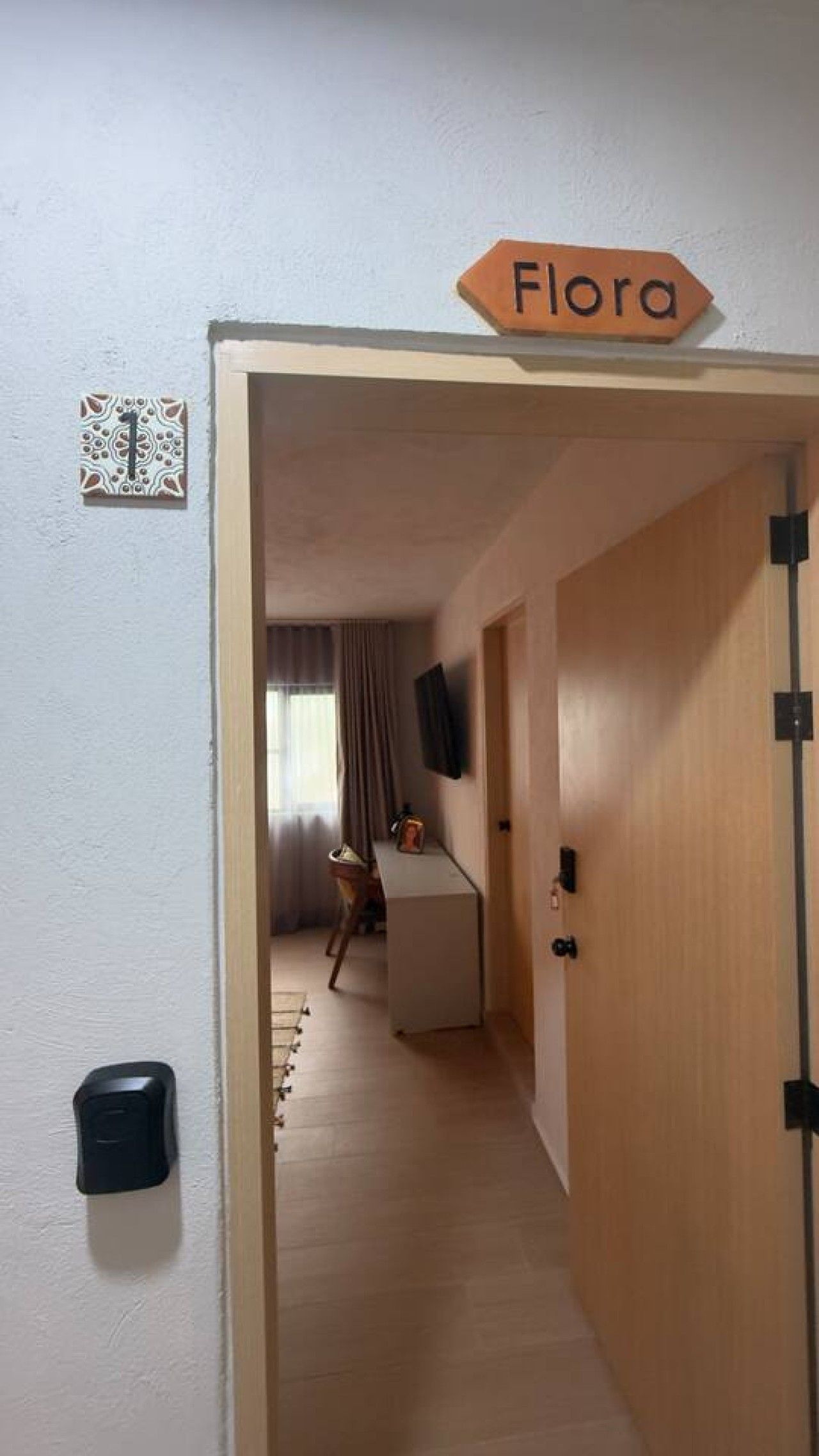 A hallway leading to a room with a door labeled 'Flora.' There is a small table and a chair in the room, and a television mounted on the wall.