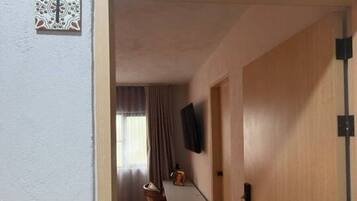 A hallway leading to a room with a door labeled 'Flora.' There is a small table and a chair in the room, and a television mounted on the wall.