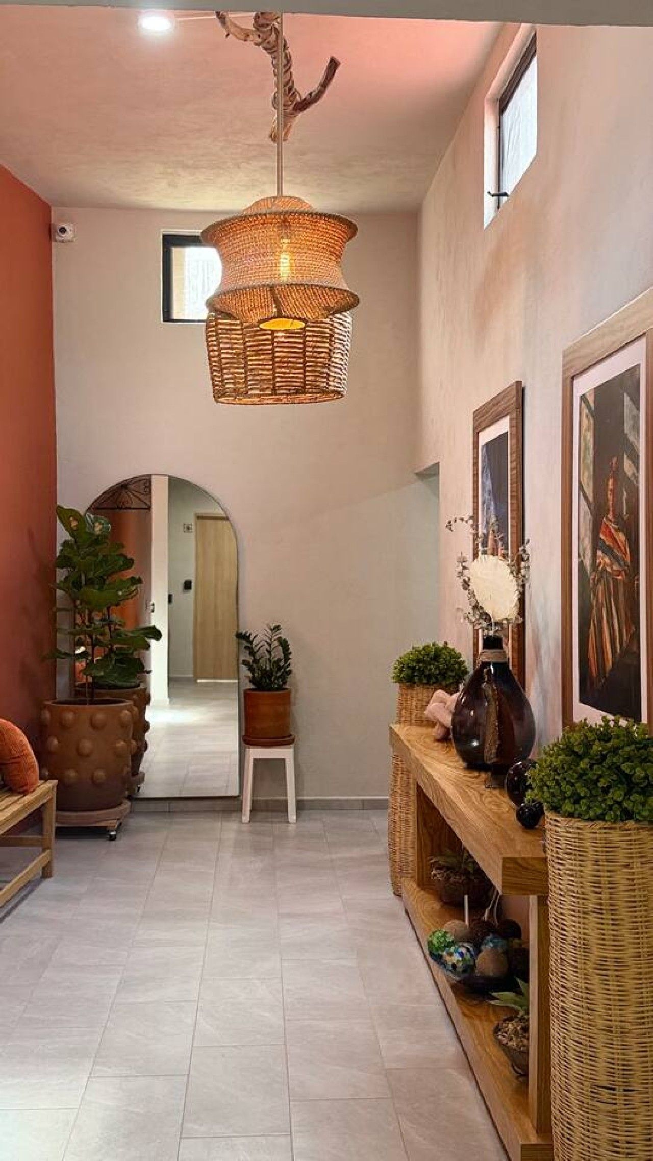 A hallway with a woven light fixture, potted plants, a wooden bench, and framed artwork on the walls.