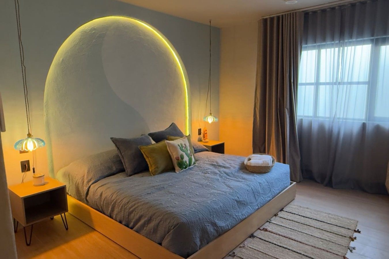 A modern bedroom with a large bed, two bedside tables, a nightstand, a wall arch, and a window with curtains.