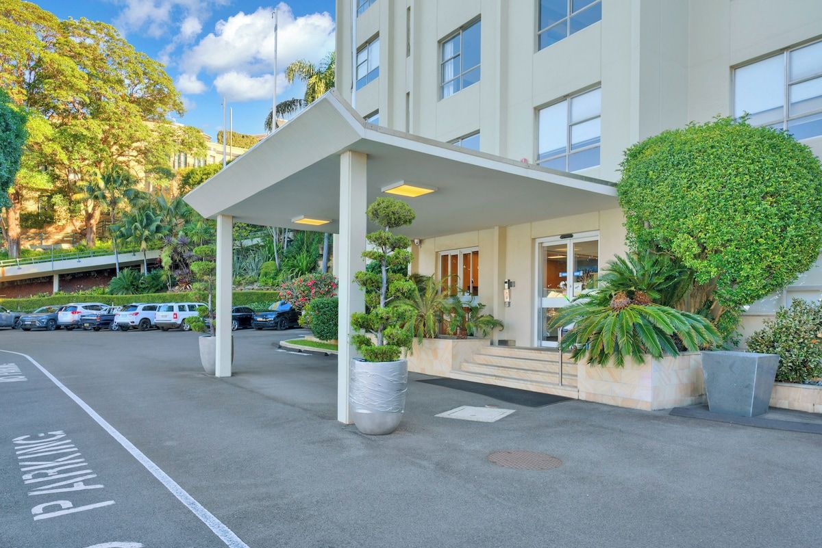 L’entrée d’un hôtel, avec un passage couvert, un parking et une végétation luxuriante.