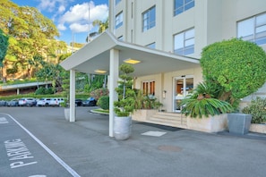 L’entrée d’un hôtel, avec un passage couvert, un parking et une végétation luxuriante.