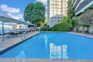 A swimming pool with a clear blue water surface, surrounded by a paved deck, a large tree, and a building in the background. - Harbourfront 1BD | Opera Views & Pool | Sydney (McMahons Point)