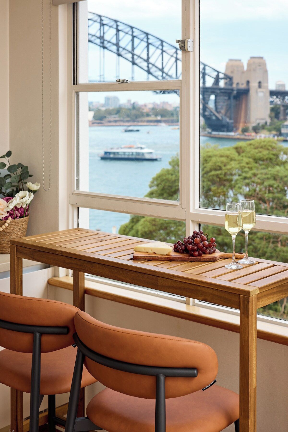 A table with a view of a bridge and water, two glasses of white wine, and a wooden board with cheese and grapes.