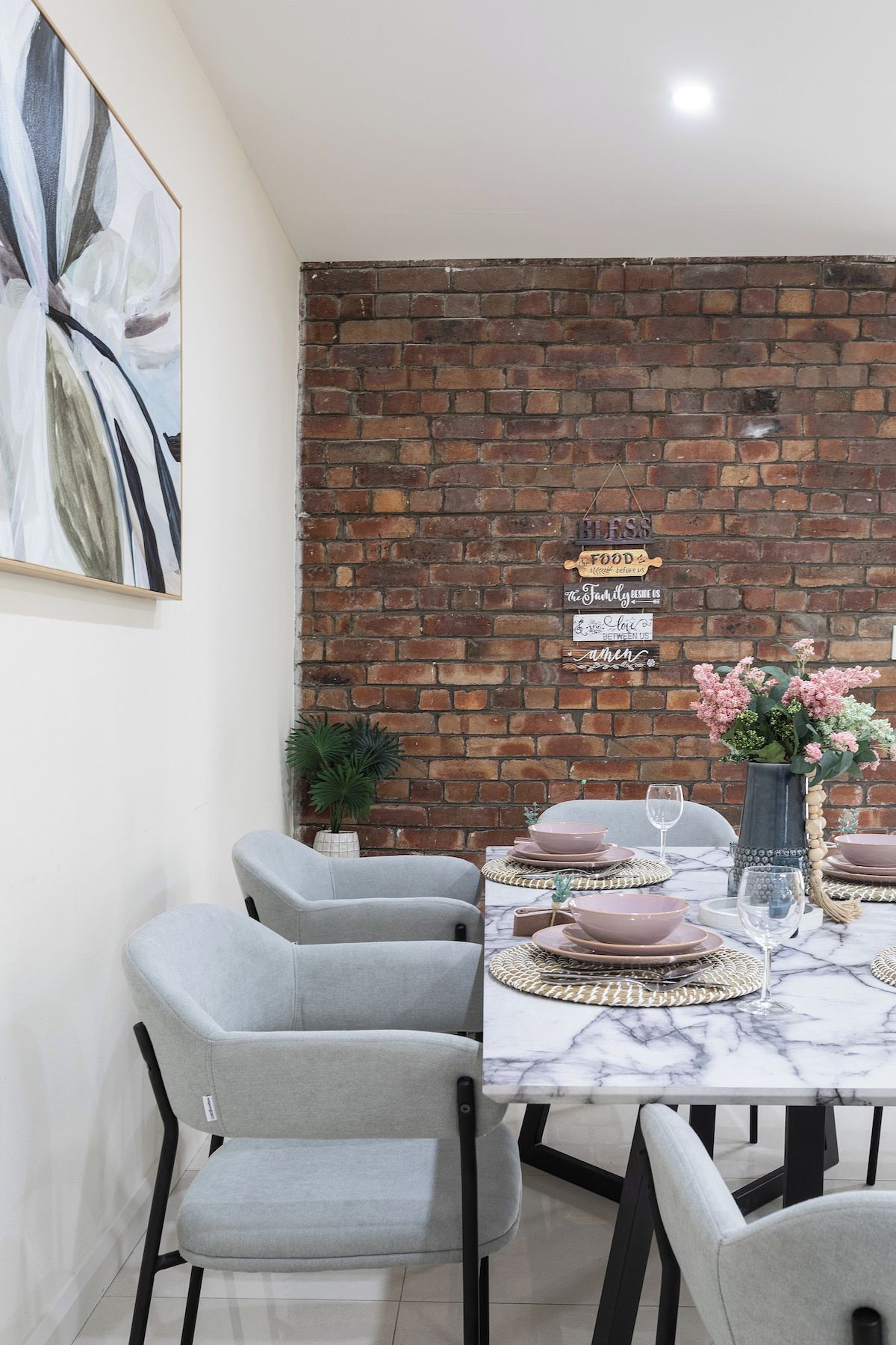 A dining area with a brick wall, a marble table, grey chairs, and a vase with flowers.