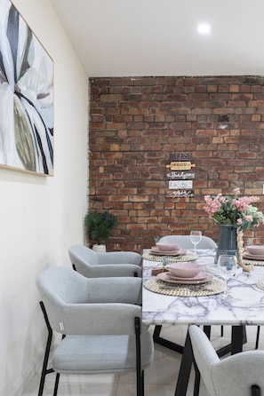 A dining area with a brick wall, a marble table, grey chairs, and a vase with flowers. - Historic 4BR Edwardian Terrace | Walk to Rail (Redfern)