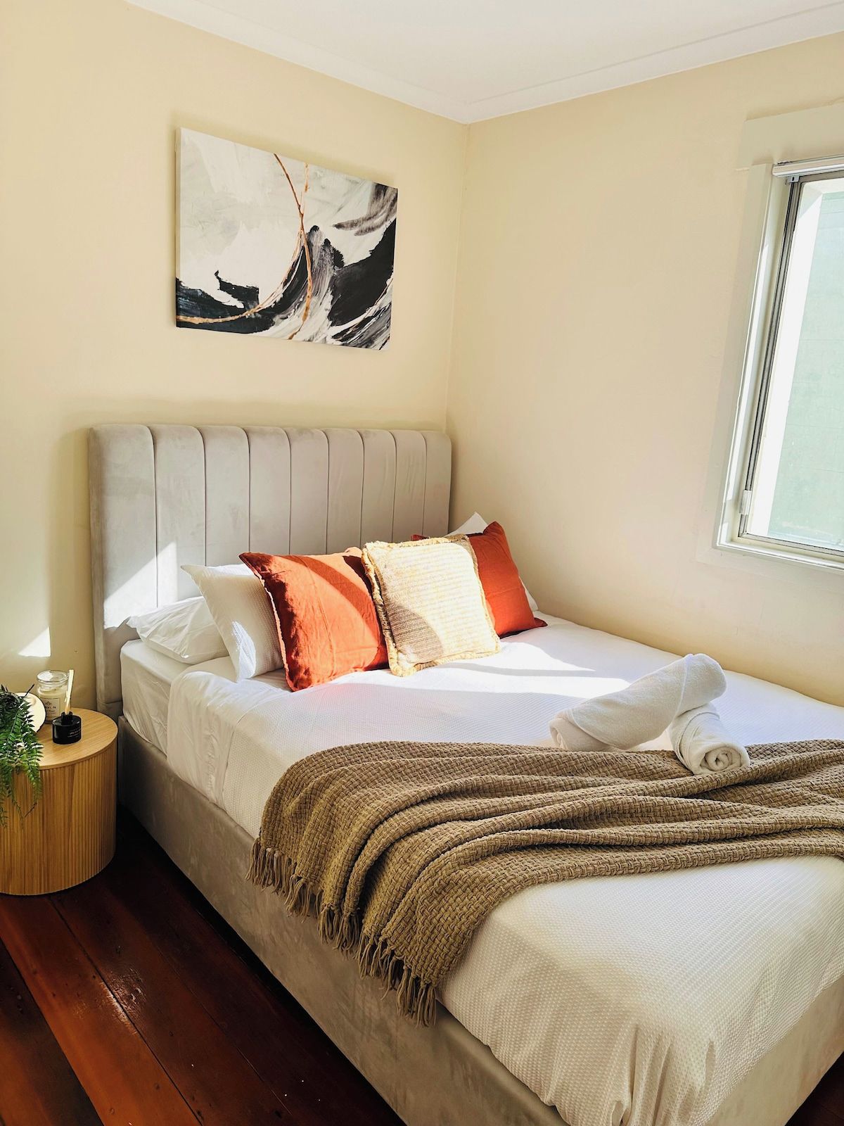 A neatly made bed with a headboard, a bedside table with a lamp, and a window with daylight.