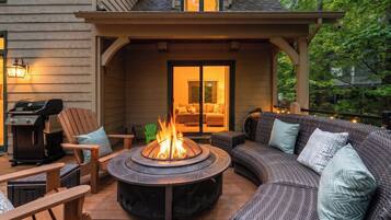 A covered outdoor seating area with a fire pit, a grill, and a sofa with cushions.