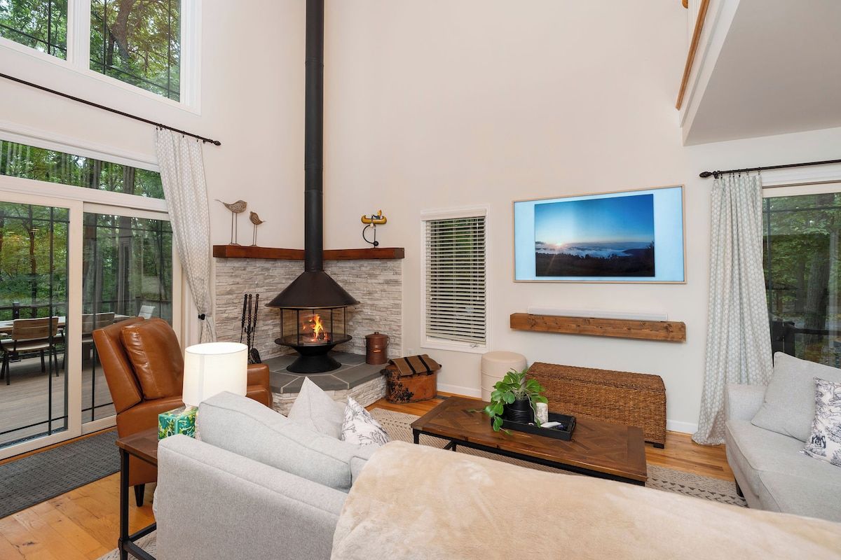 A cozy living room with a fireplace, comfortable seating, and a large window overlooking a forest.