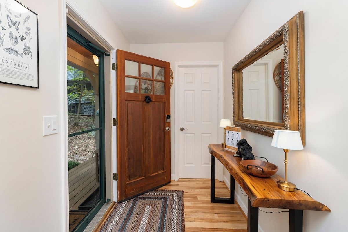 A hallway with a wooden door, a white door, a mirror, a lamp, and a small table with a bowl and a figurine.