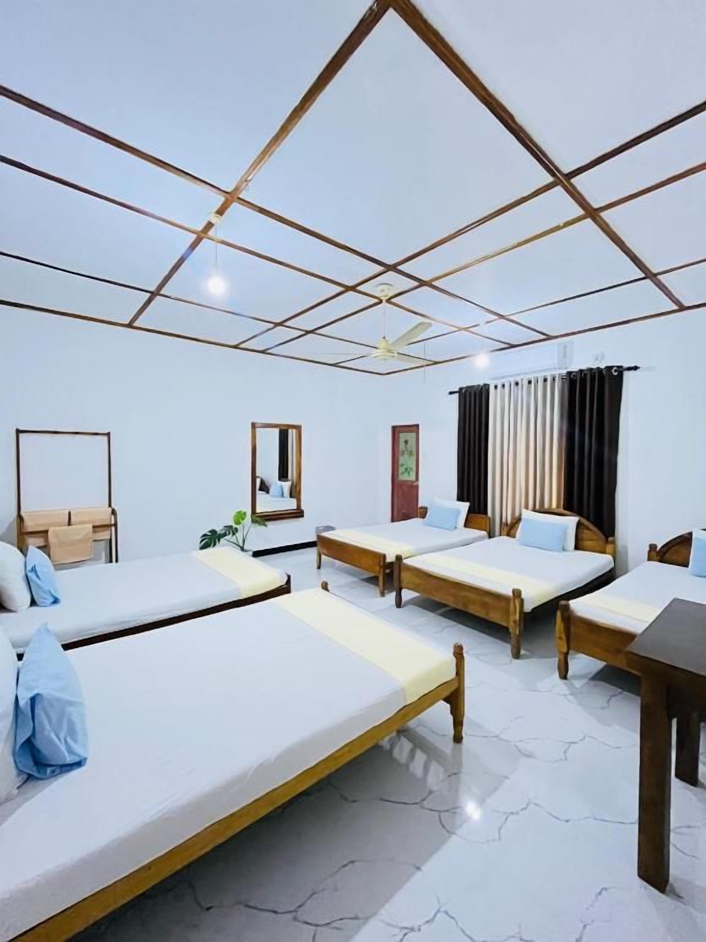 A hotel room with four single beds, a wooden table, a ceiling fan, and a large window with curtains.