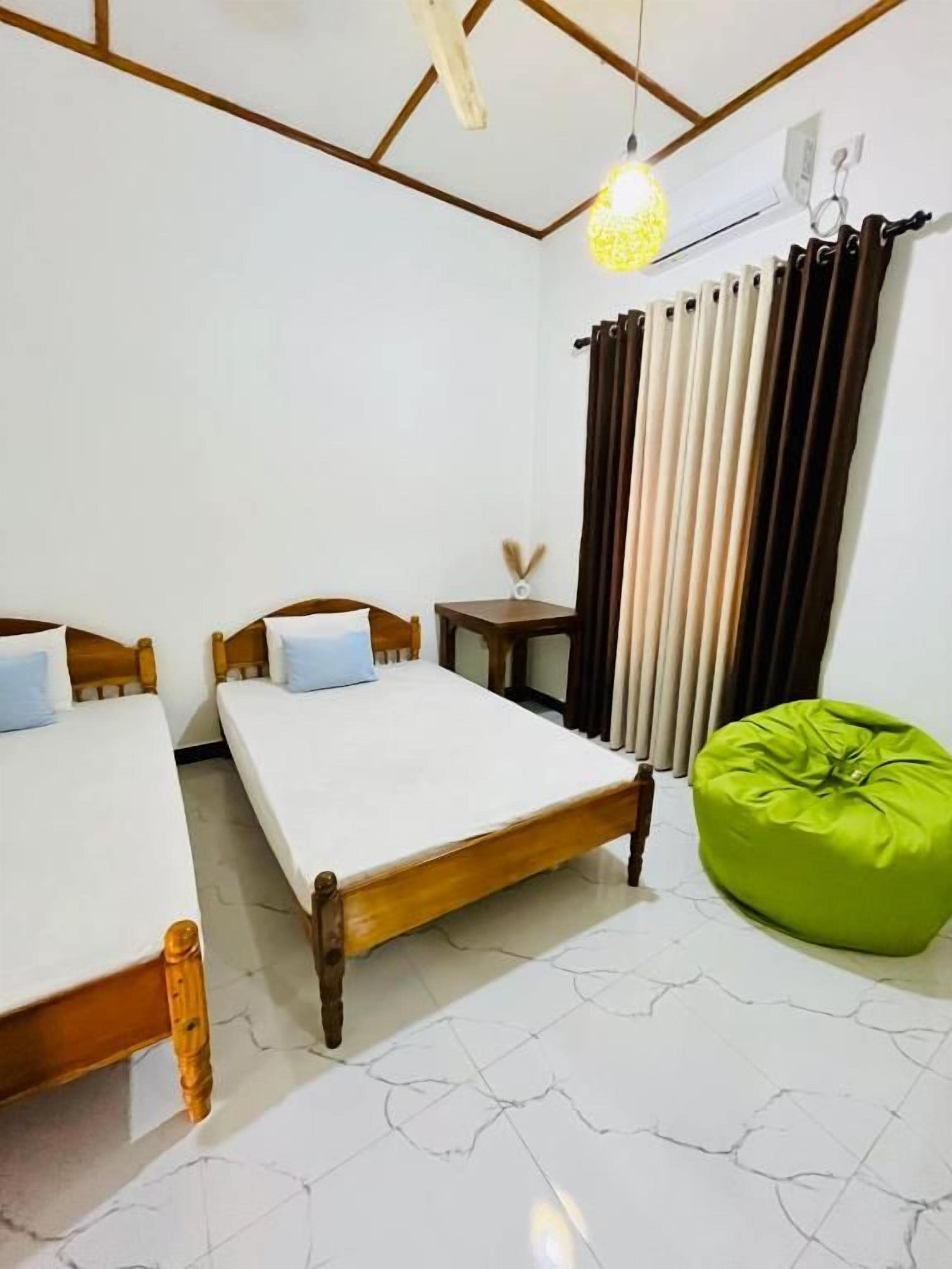 A room with two beds, a green bean bag, a small table, and a hanging light fixture.