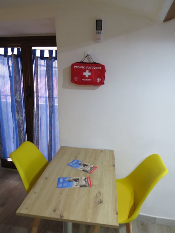 Habitació amb taula de fusta, dues cadires grogues, un kit de primers auxilis vermell i una finestra amb cortines blaves.
