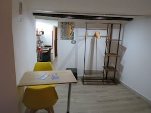 Una zona de menjador amb taula i cadires, una cuina al fons i un moble prestatges de fusta.