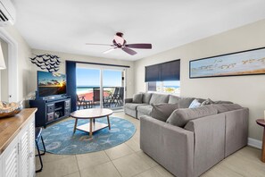 A living room with a sofa, a TV, a ceiling fan, and a view of the ocean. - Regency Towers 808 W (Pensacola Beach)