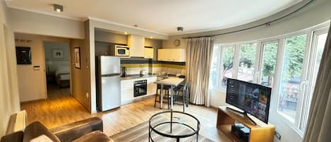 A modern living room with a sofa, a coffee table, and a TV. There is a kitchen with a refrigerator, microwave, and stove. The room has large windows with curtains.
