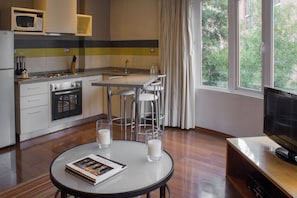A modern kitchen with a microwave, stove, and bar stools. There is a dining table with a book and glasses. A TV is on a stand to the right. - Luxury 1-Bedroom Apartment in Lastarria (Santiago)