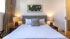 Un lit bien fait, avec du linge de lit blanc, une tête de lit grise et un coussin décoratif. Deux lampes de chevet sont posées sur des tables de chevet en bois, et une œuvre d’art est accrochée au mur au-dessus du lit.