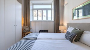 A hotel room with two beds, a window with curtains, a chair with a patterned cushion, and a wall-mounted lamp. - Design 2BR Duplex with AC by LovelyStay (Lisboa)