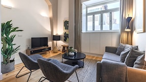 Un salon moderne doté d’un téléviseur à écran plat, de deux fauteuils, d’un canapé, d’une table basse et d’une grande fenêtre agrémentée de rideaux.
