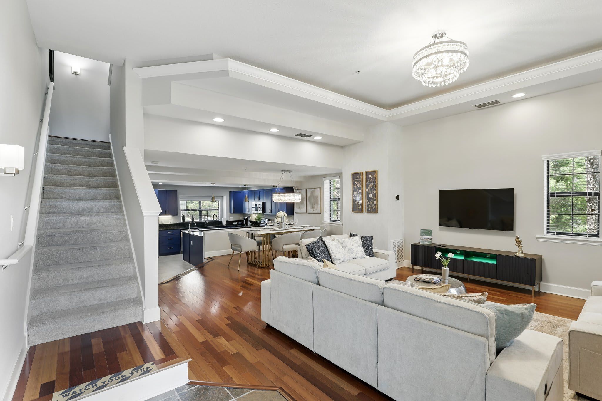 Una sala de estar moderna con un sofá gris, un televisor de pantalla plana y una escalera que lleva a una zona de cocina.