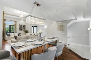 A modern dining area with a long table set for six, a chandelier, and a sofa in the background. - Magical Townhome Near Disney w Resort Pool Access (Orlando)