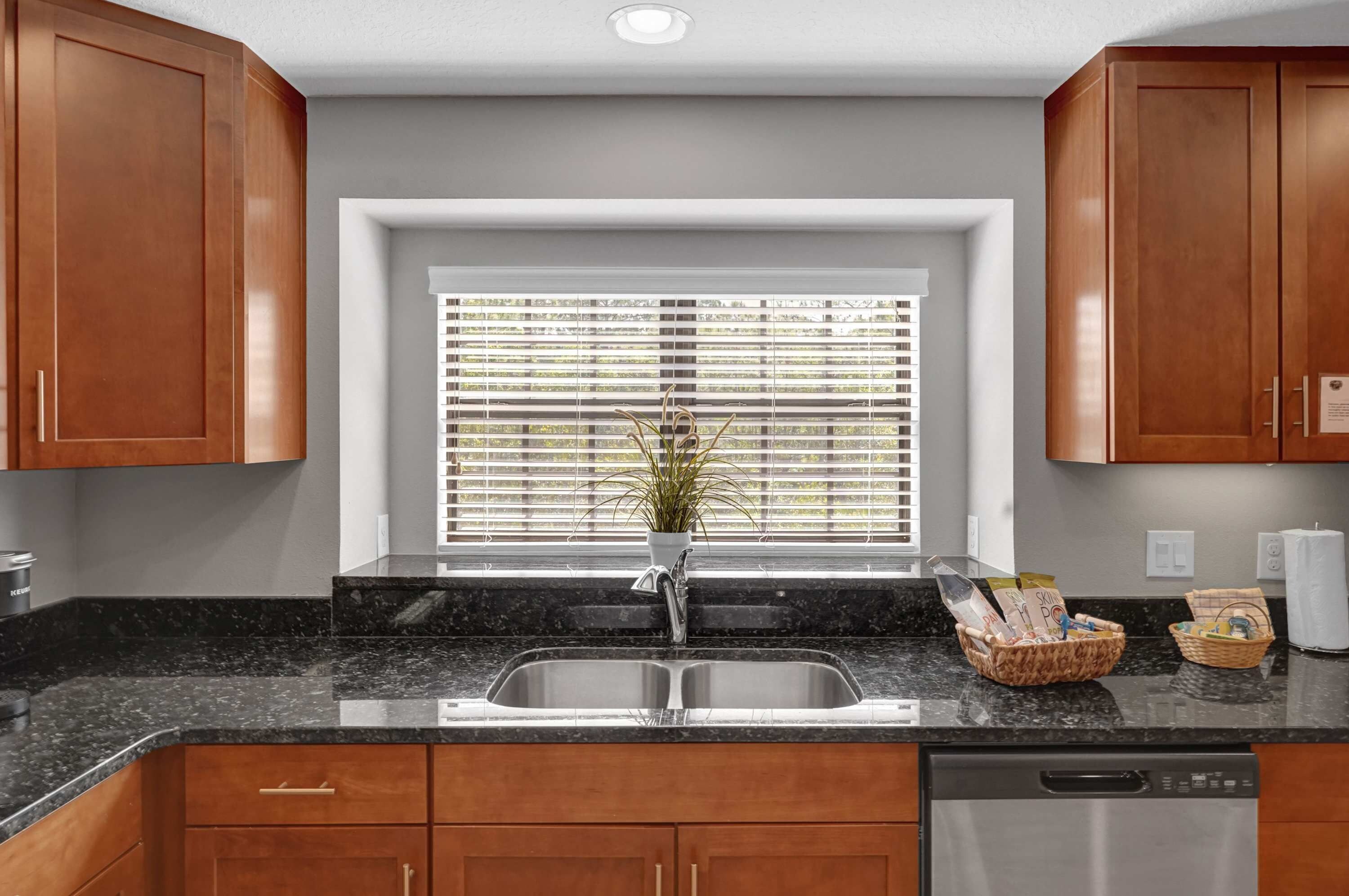 A kitchen with wooden cabinets, a granite countertop, a stainless steel sink, and a window with blinds.